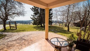 Un porche cubierto con suelo de baldosas, una mesa de mimbre, una hamaca y vistas a un paisaje con césped y árboles.
