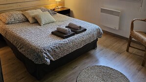 Un dormitorio con cabecera de madera, cama con colcha estampada, dos almohadas, lámpara de noche, cuadro enmarcado en la pared, ventana con persianas y una silla de mimbre.