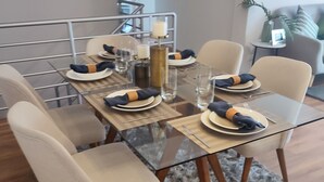 Un coin repas avec une table en verre dressée pour quatre personnes, entourée de chaises. Un tapis moelleux recouvre le sol, et un escalier est visible au fond.