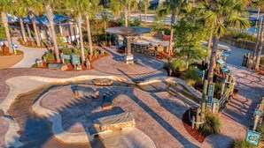 A water-themed park area with a central fountain, surrounded by palm trees and a paved walkway.