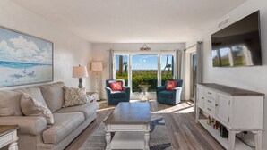 A spacious living room with a beige sofa, two blue armchairs, a coffee table, and a TV mounted on the wall.