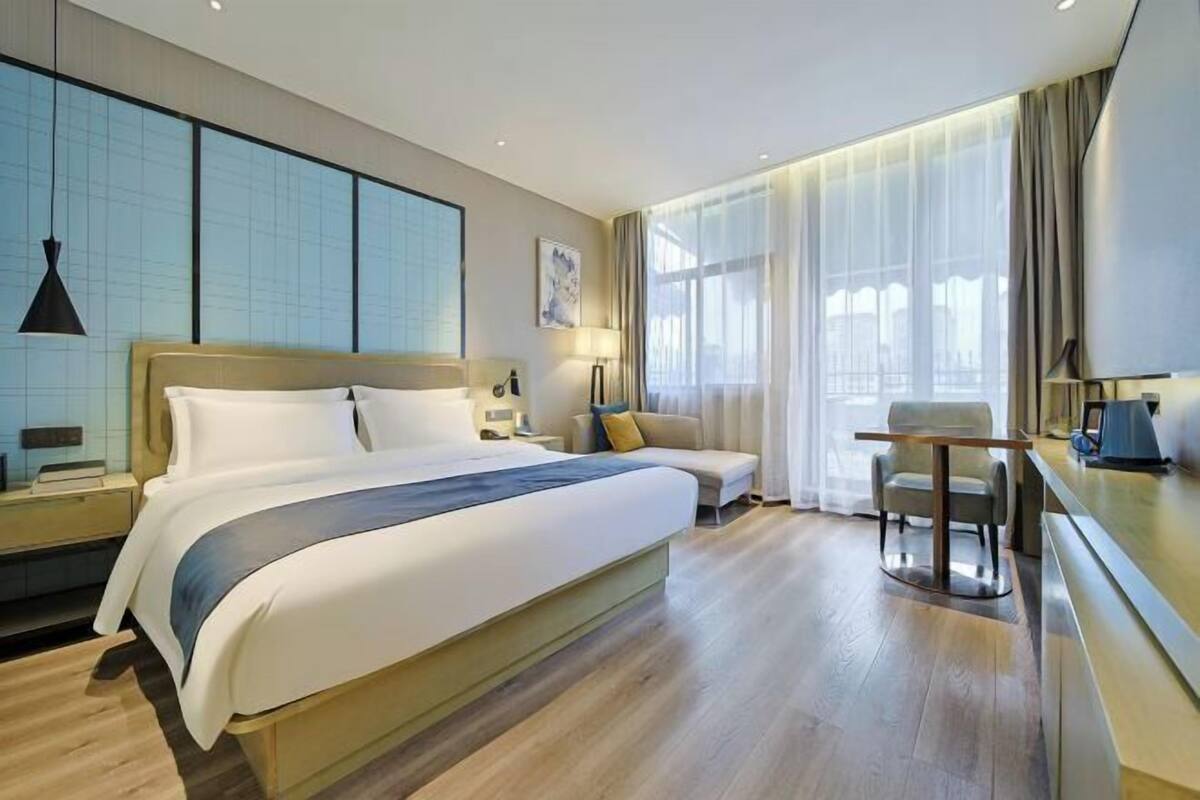 Quarto de hotel com uma cama grande, uma mesa, uma cadeira e uma janela com cortinas.