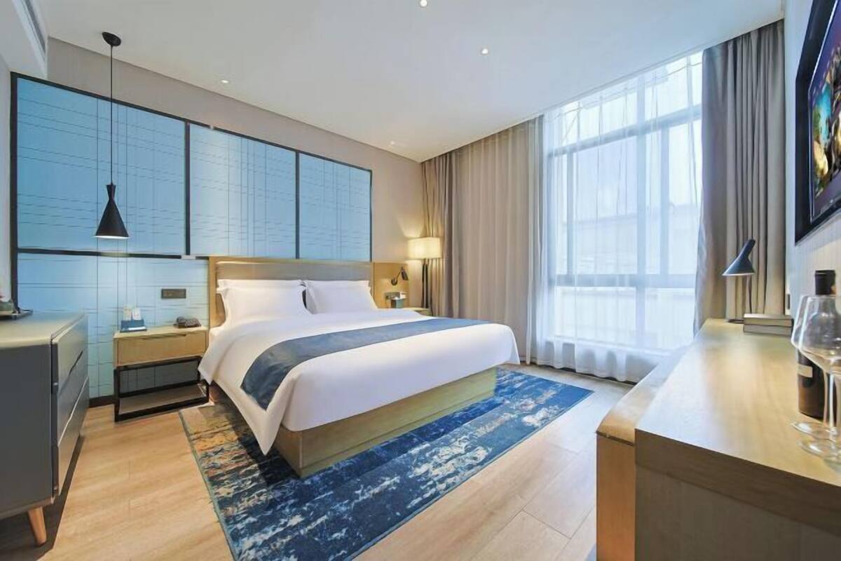 Quarto de hotel moderno com uma cama grande, uma escrivaninha, mesa de cabeceira, televisão na parede e uma janela ampla com cortinas.