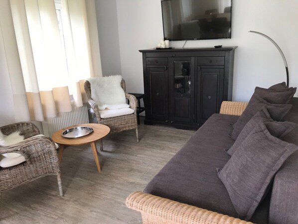 A living room with a dark brown sofa, two wicker chairs, a small round table, a dark wooden cabinet, and a flat-screen TV mounted on the wall.