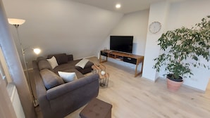 Uma sala de estar moderna com um sofá seccional, uma TV sobre um suporte de madeira, uma planta em vaso e um relógio na parede.