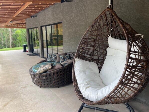 A hanging chair with a white cushion and a wicker ottoman with patterned cushions.