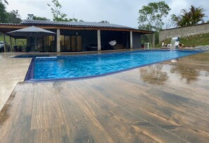 A swimming pool with a wooden deck, a covered seating area, and a building with a tiled roof.