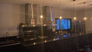 Une cuisine moderne dotée d’appareils électroménagers en acier inoxydable, d’un éclairage suspendu et d’une grande fenêtre offrant une vue sur une forêt.