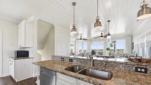 A kitchen with granite countertops, white cabinetry, stainless steel appliances, pendant lighting, ceiling fans, and a view to the outdoors. - Blue Pearl #106-Soundfront with Private Dock, Kayaks & Walkable Beach Access! (North Topsail Beach)