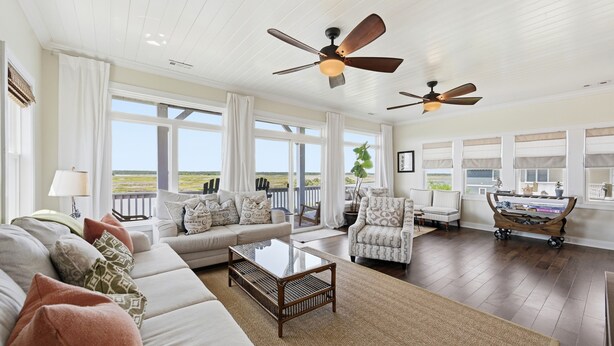 A spacious living room with a large window, a ceiling fan, a sofa, a coffee table, and a bookshelf. - Blue Pearl #106-Soundfront with Private Dock, Kayaks & Walkable Beach Access! (North Topsail Beach)