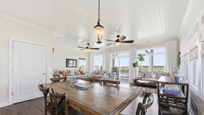 A spacious dining area with a wooden table, chairs, ceiling fans, and large windows offering a view. - Blue Pearl #106-Soundfront with Private Dock, Kayaks & Walkable Beach Access! (North Topsail Beach)