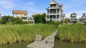 A wooden dock leading to a house with a balcony, surrounded by tall grass. - Blue Pearl #106-Soundfront with Private Dock, Kayaks & Walkable Beach Access! (North Topsail Beach)