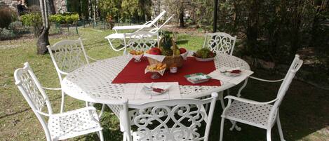 A round white metal table with a red tablecloth, surrounded by white metal chairs, set outdoors with a stone building and greenery in the background.