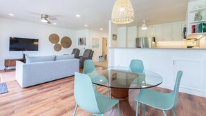 A modern living room with a glass-top dining table, teal chairs, a grey sofa, and a ceiling fan. - Hawaii Life Rental Presents Emmalani 423 (Princeville)