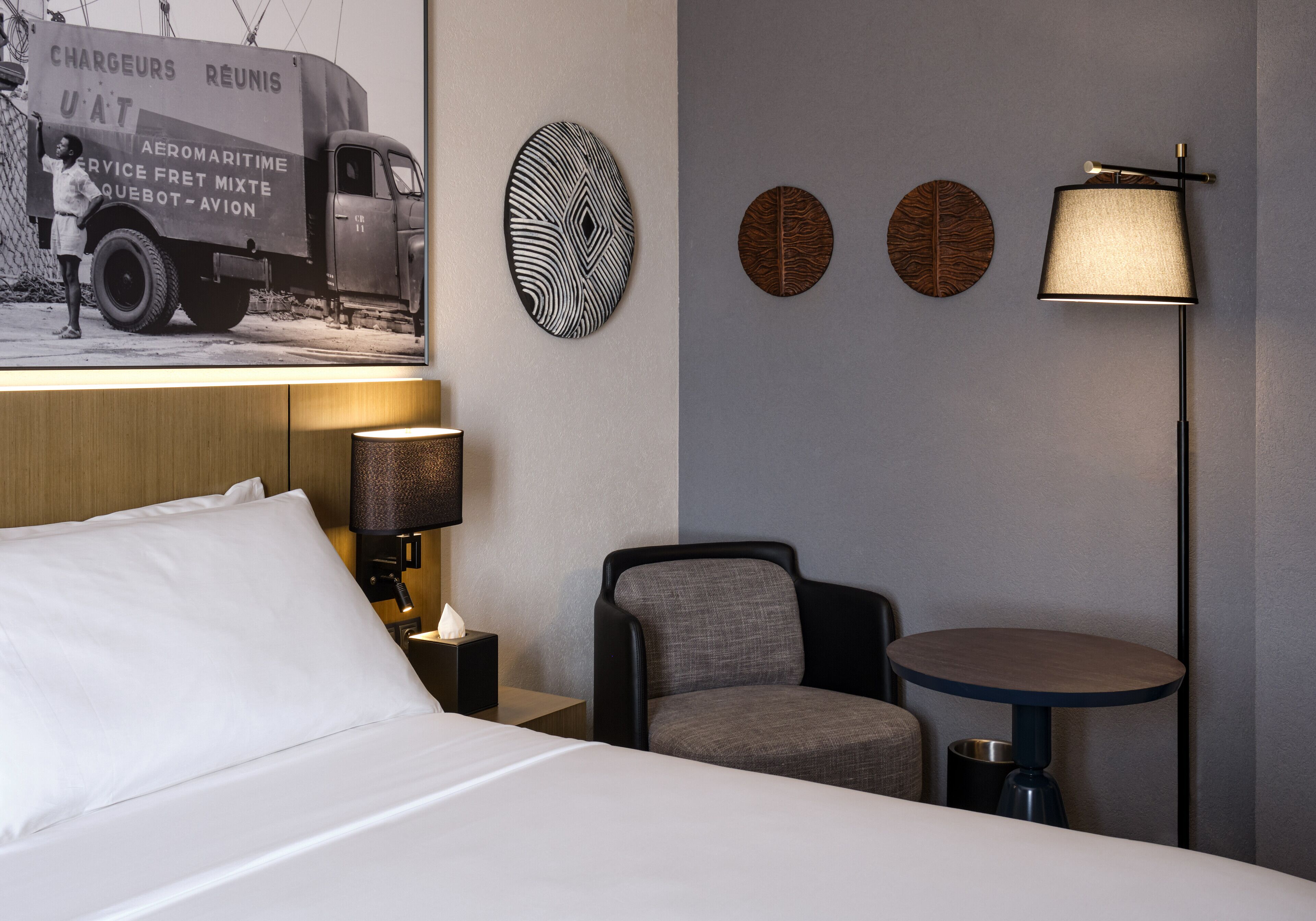 A hotel room with a bed, a chair, a small table, a lamp, and wall art including a truck and leaf patterns.