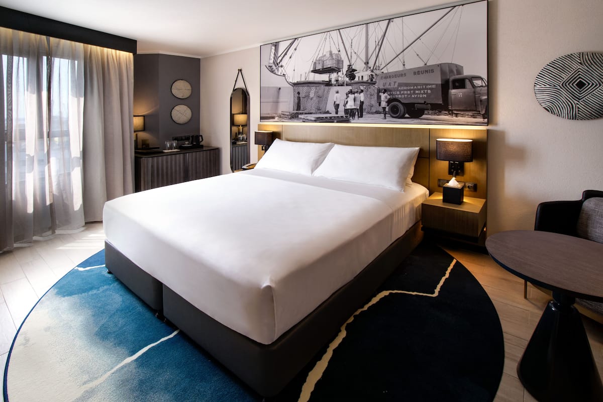 Habitación de hotel con una cama grande, un escritorio, una silla, una lámpara de escritorio, un reloj de pared y un cuadro enmarcado.