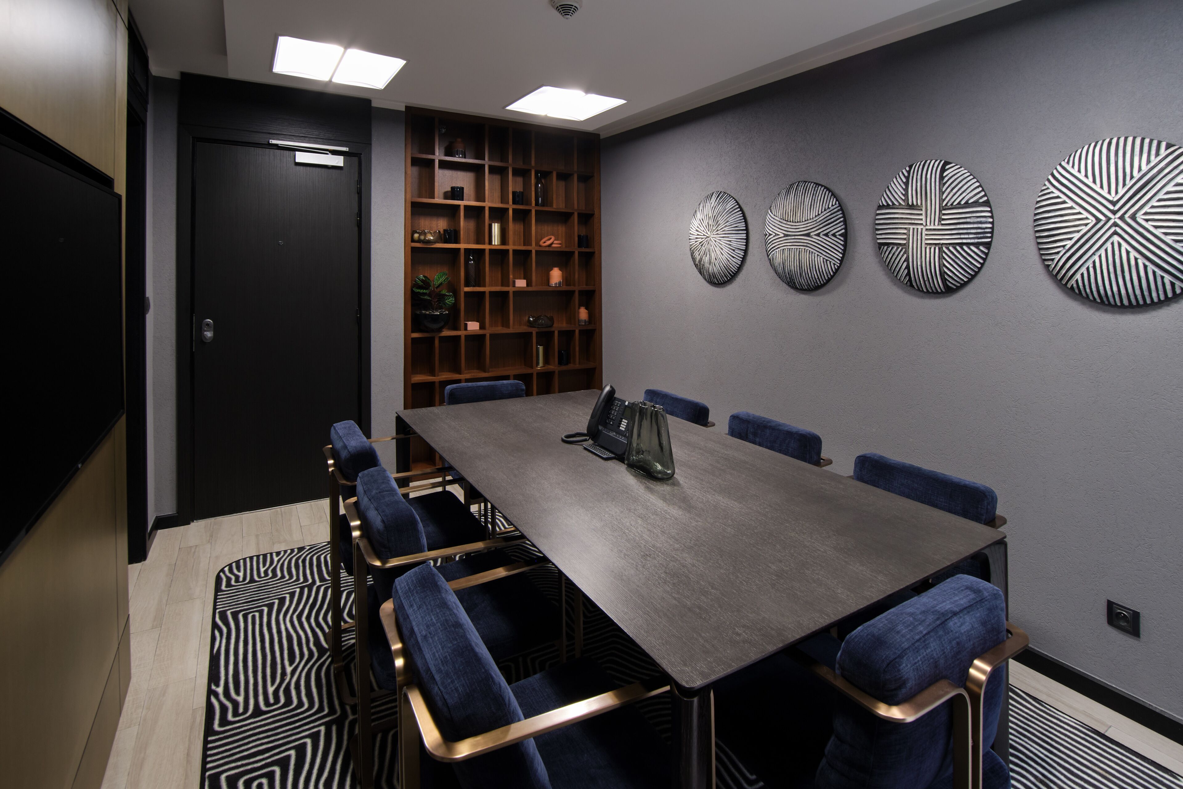 Een congreszaal met een lange tafel, blauwe fluweelstoelen, een boekenkast en decoratieve muurkunst.