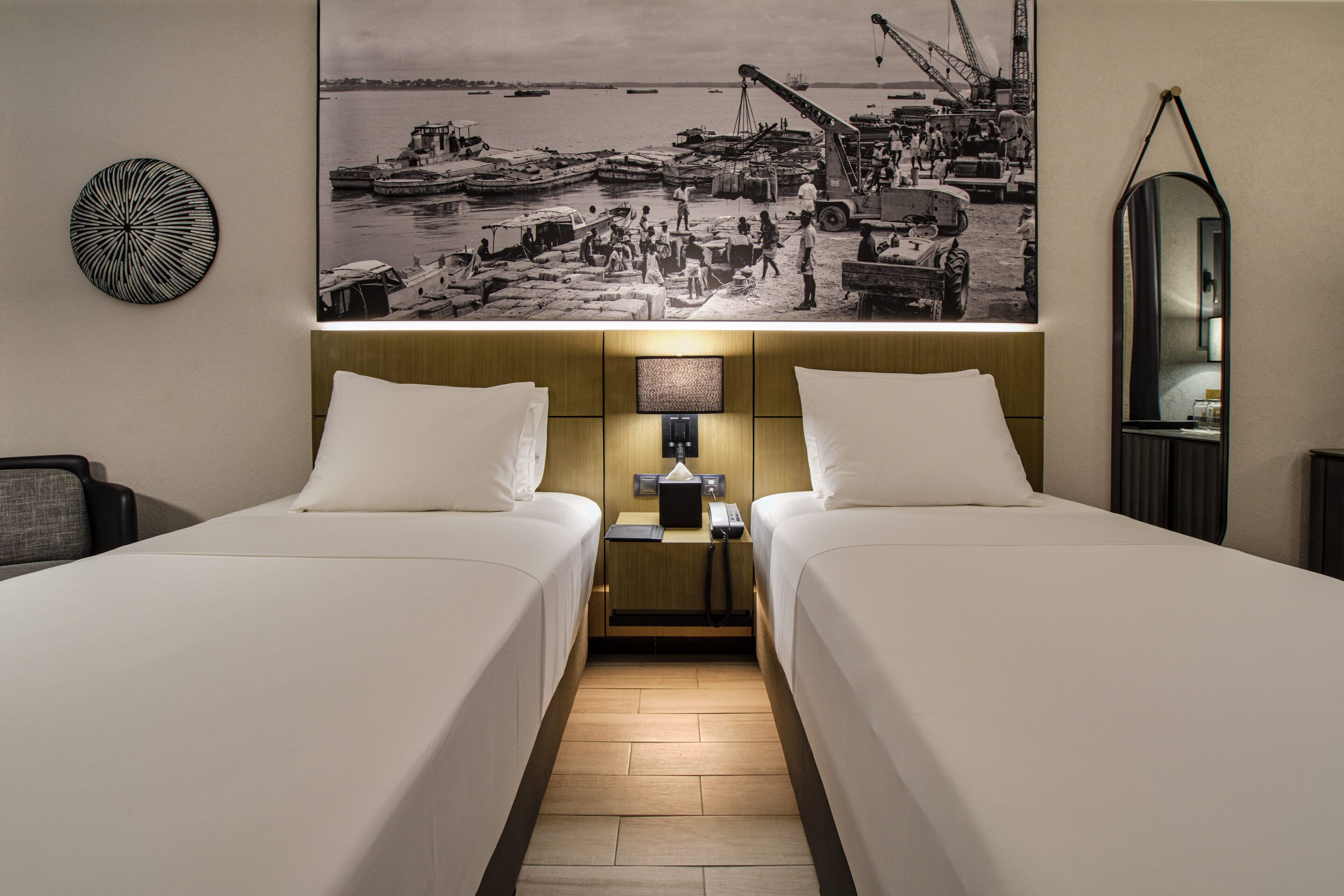 Habitación de hotel con dos camas, una mesita de noche con lámpara, un televisor y una fotografía grande en blanco y negro en la pared.