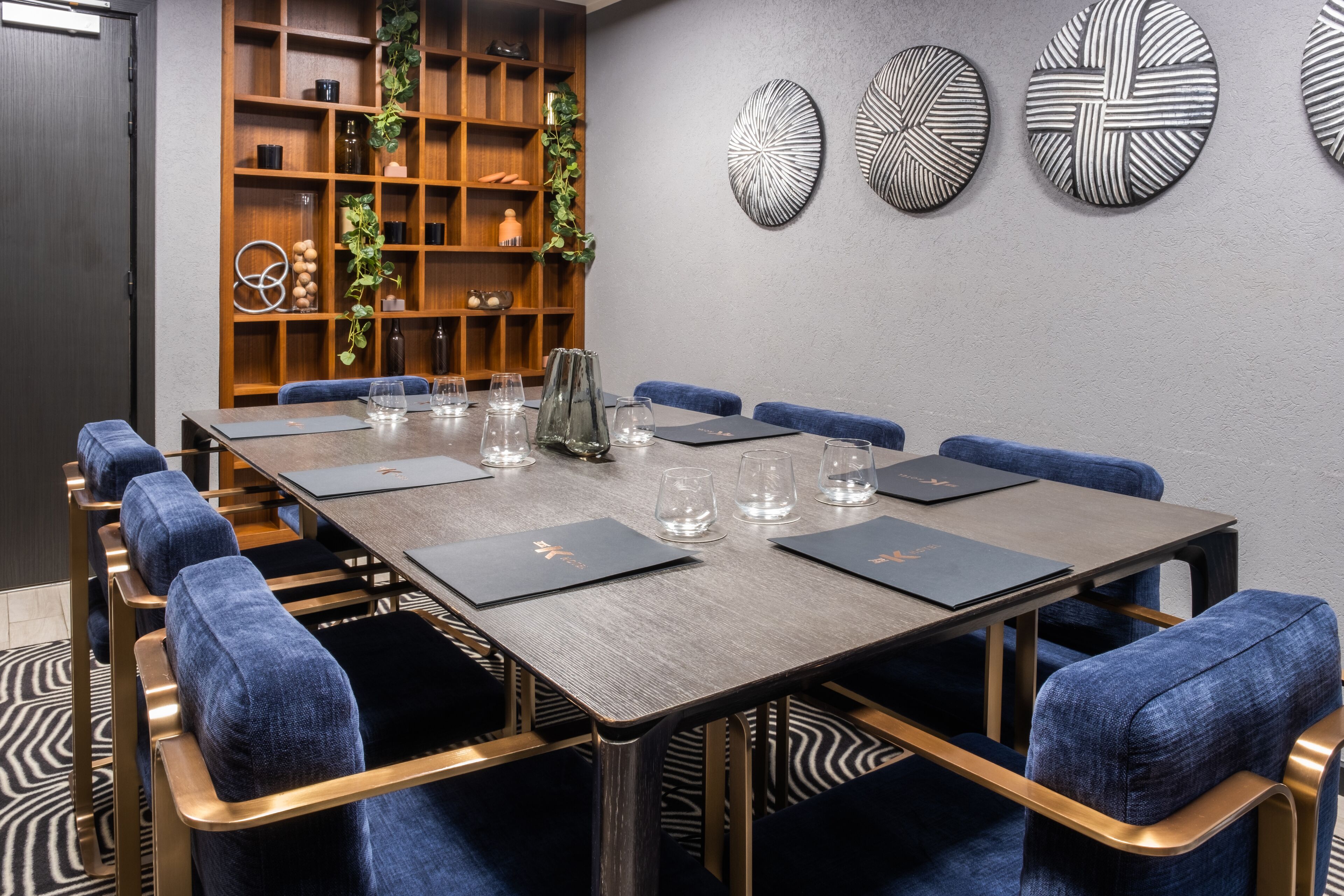A conference room with a long table, blue upholstered chairs, and a wooden bookshelf with decorative items.