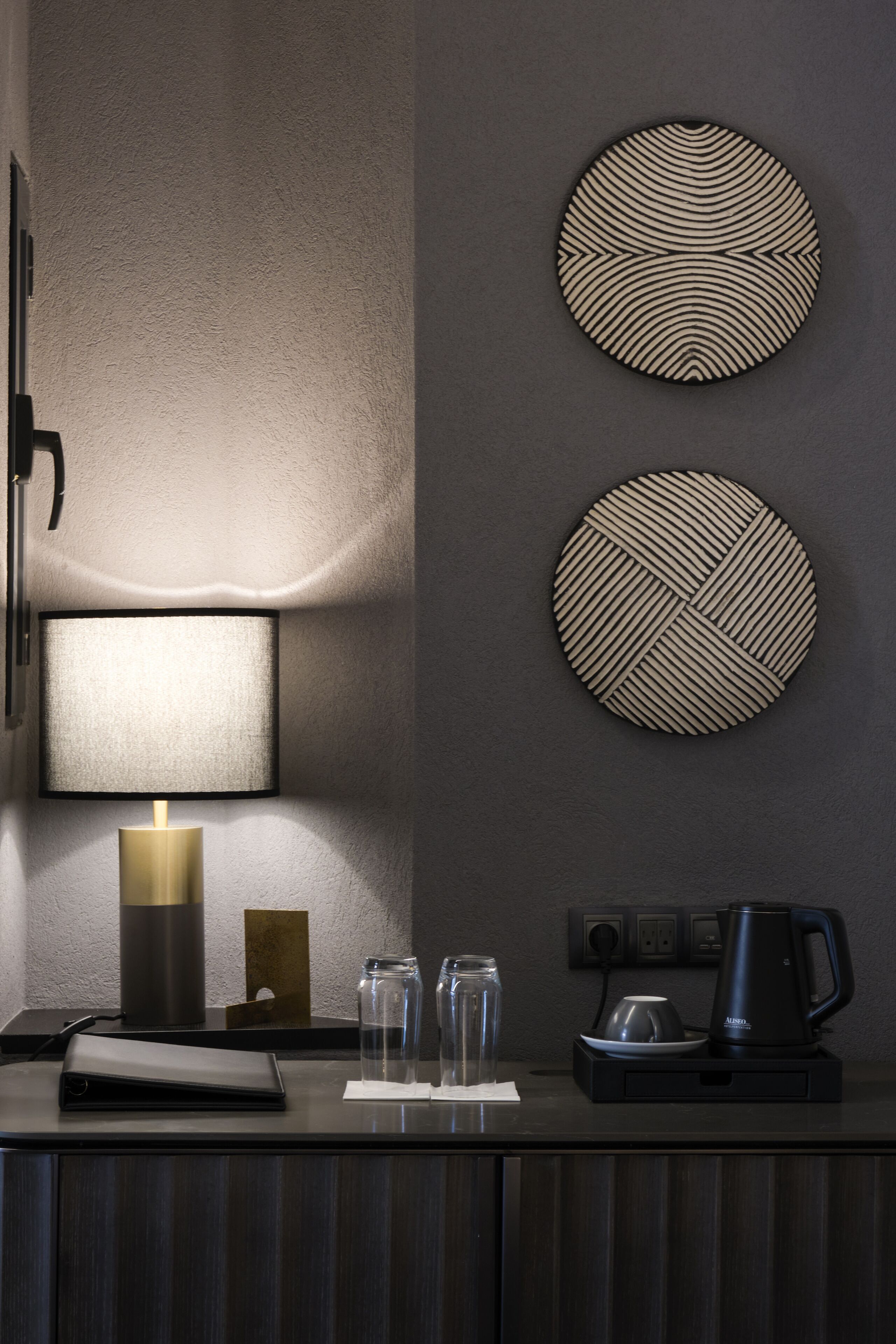 Habitación de hotel moderna con detalles en madera oscura, una lámpara de pared, dos piezas de arte circular, una cafetera y una pequeña mesa con un cuaderno y vasos.