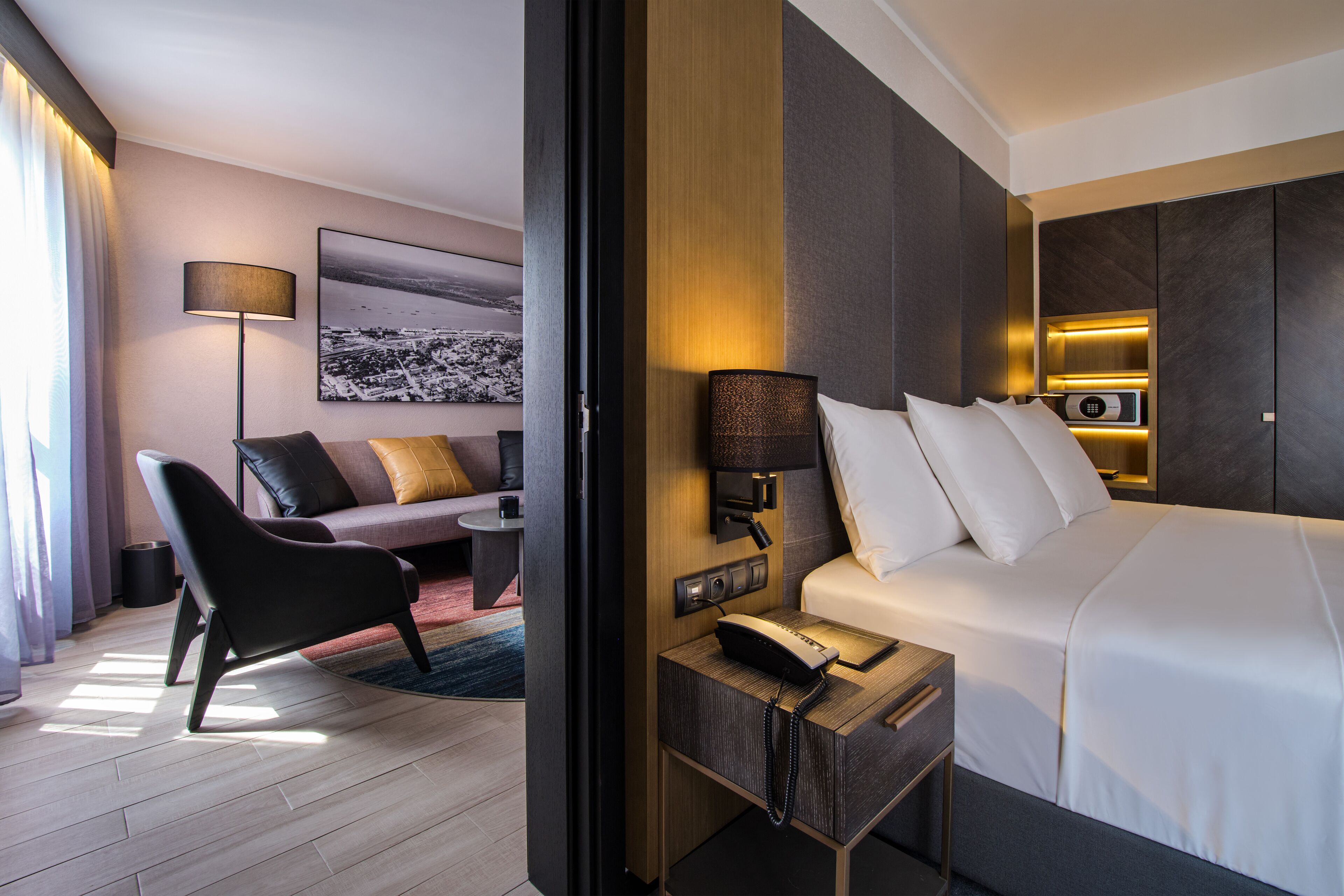 A modern hotel room with a neatly made bed, a bedside table with a lamp, a chair, and a sofa.