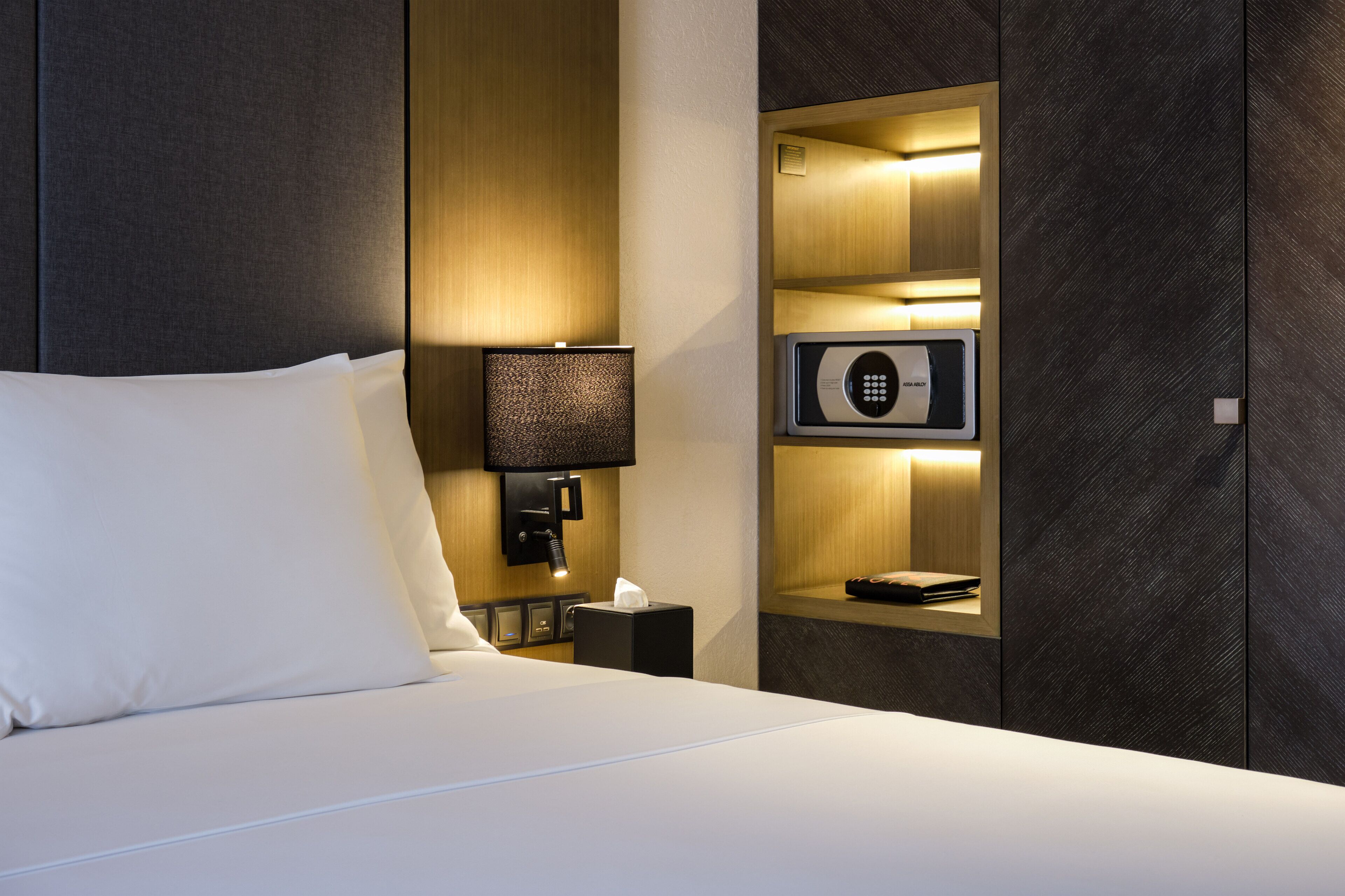 Quarto de hotel com cama bem arrumada, luminária de cabeceira, minibar e cofre embutido.