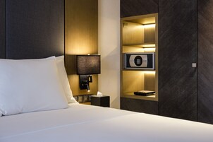 Habitación de hotel con una cama bien hecha, una lámpara de noche, un minibar y una caja fuerte empotrada.