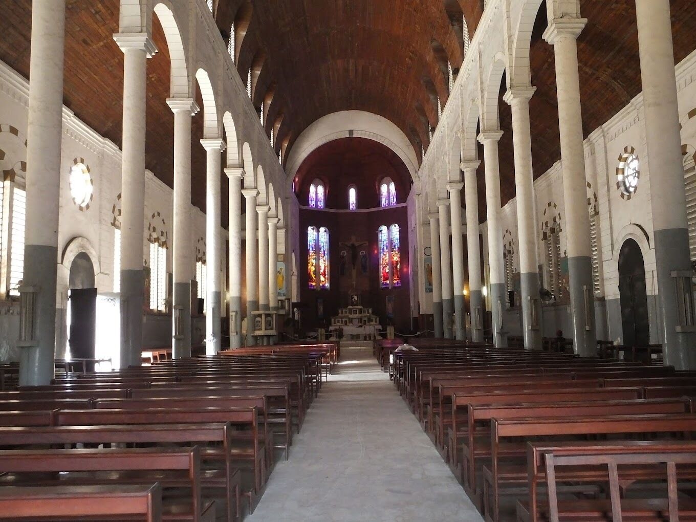 A church interior with rows of wooden pews, white columns, and stained glass windows.