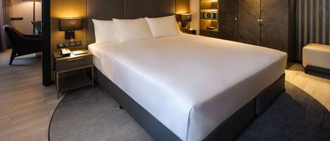 A modern hotel room with a large bed, a desk with a lamp, a chair, and a wardrobe.