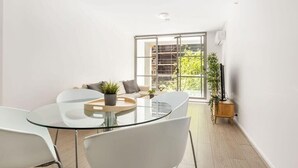 Una sala de estar moderna con una mesa de comedor de vidrio, sillas blancas, un sofá y un ventanal con vistas a edificios y vegetación.