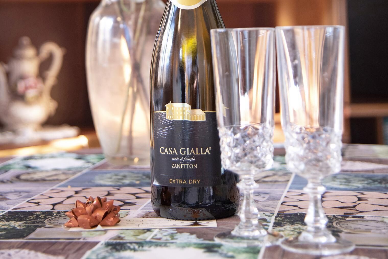 Una botella de vino Casa Gialla Extra Dry Zannetton, dos copas y un lazo decorativo sobre una mesa de baldosas.