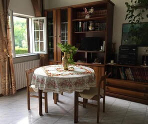 Ein Essbereich mit einem runden Tisch, bedeckt mit einer blumigen Tischdecke, umgeben von Holzsesseln. Auf dem Tisch steht eine Vase mit einer Pflanze.