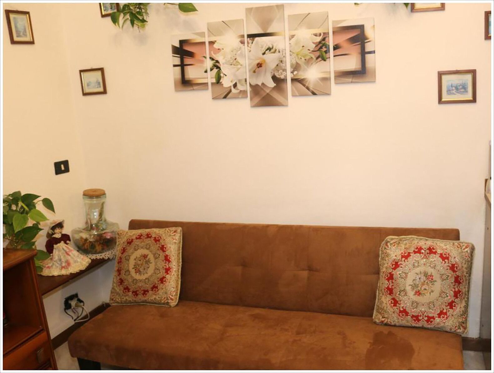 A brown sofa with two decorative pillows, a small table with a plant and a figurine, and a wall with framed pictures and a floral arrangement.