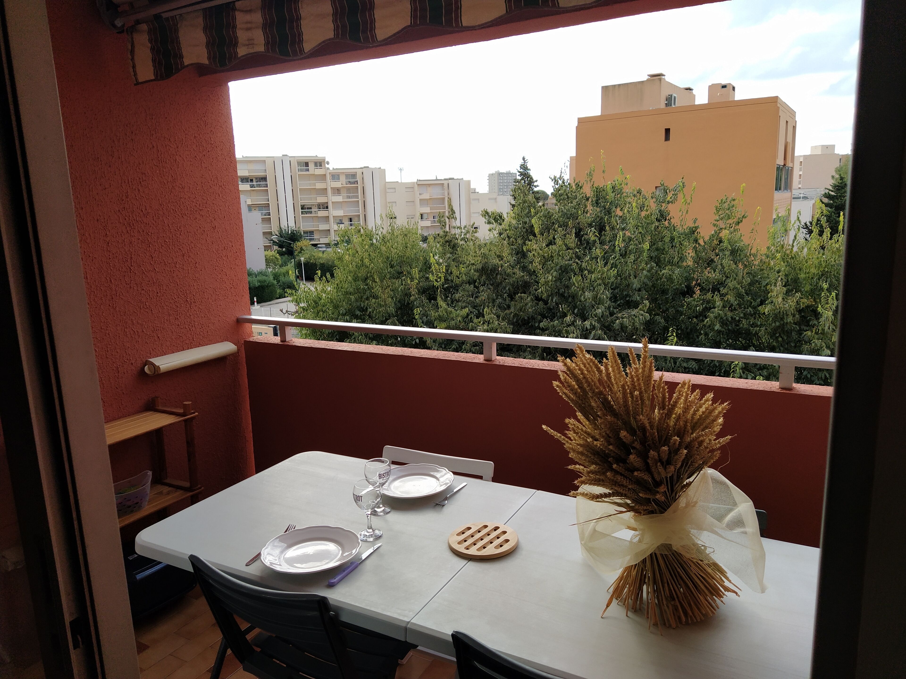 A balcony with a table set for two, a vase of dried plants, and a view of buildings and greenery.