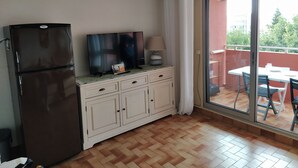 Eine kompakte Küche mit Kühlschrank, einem Flachbildfernseher, einem kleinen Tisch mit Lampe und einer Glastür, die zu einem Außenbereich mit Tisch und Stühlen führt.