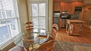 Un comedor con mesa de vidrio y sillas de madera, una cocina con armarios y electrodomésticos de madera, y vistas a través del ventanal.