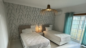 A modern bedroom with two beds, a large window with curtains, and a patterned wall. - Villa 3 Irmãos (Alvor)
