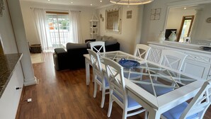 A modern dining area with a glass-top table and white chairs, a chandelier, and a view of the outdoors through large windows. - Villa 3 Irmãos (Alvor)