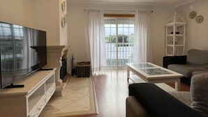 A modern living room with a flat-screen TV, a sofa, a coffee table, and a balcony with a view. - Villa 3 Irmãos (Alvor)