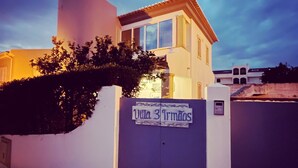 A building with a sign reading "Villa 3 Trindos" at the entrance, a cobblestone pathway, and a clear sky. - Villa 3 Irmãos (Alvor)