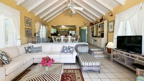 A spacious living room with a sofa, ottoman, and a TV on a stand. There is a dining area in the background with chairs and a table.