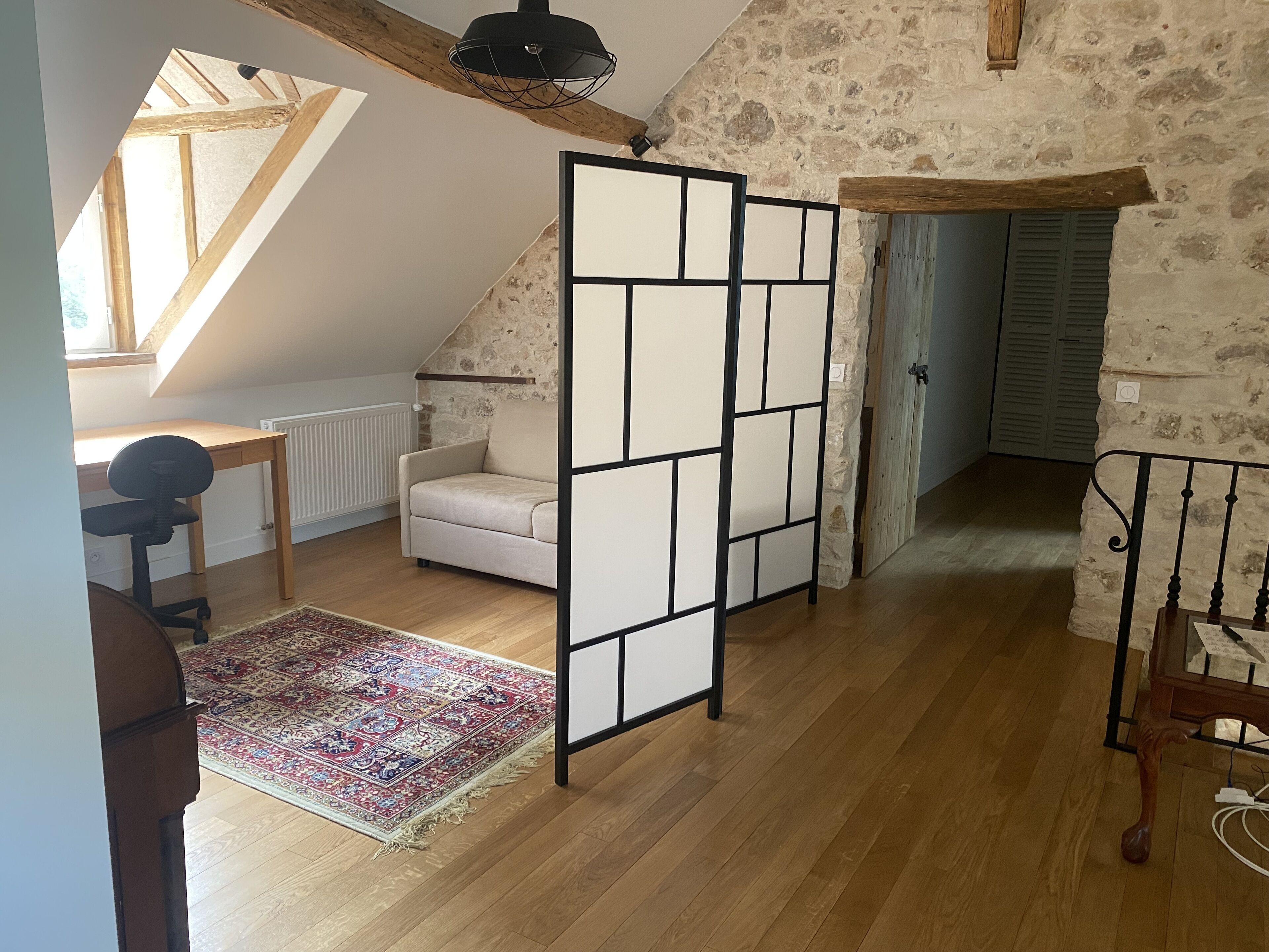 A room with a sloped ceiling, a wooden desk, a chair, a sofa, a patterned rug, and a decorative partition.