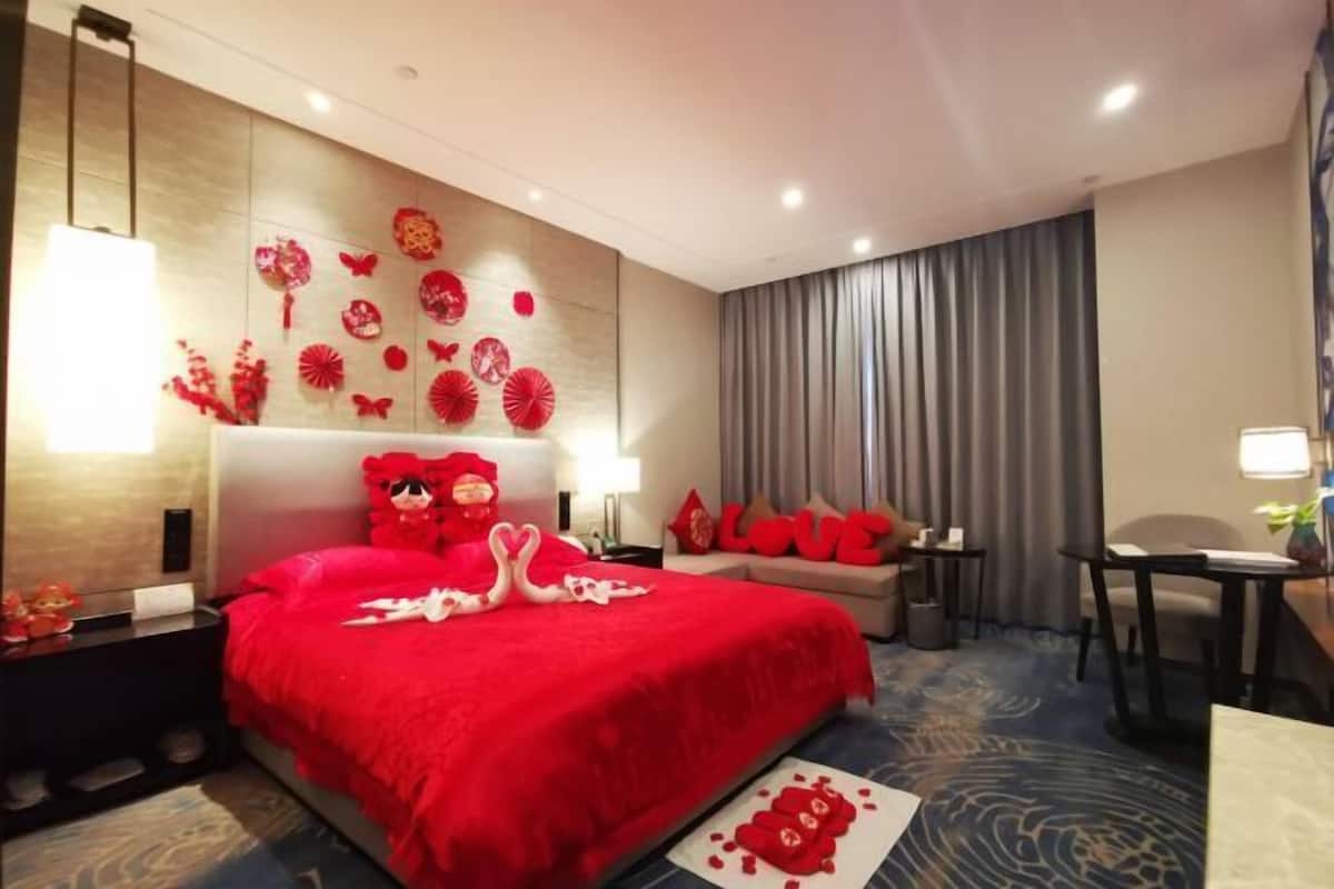 A hotel room with a red bedspread, a headboard with red decorations, a small table, a chair, and a sofa.
