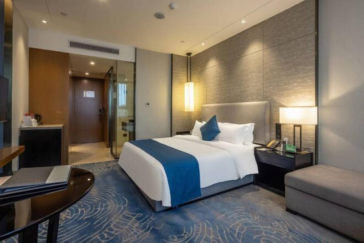 A modern hotel room with a large bed, a bedside table with a lamp, a desk with a chair, and a glass partition wall.
