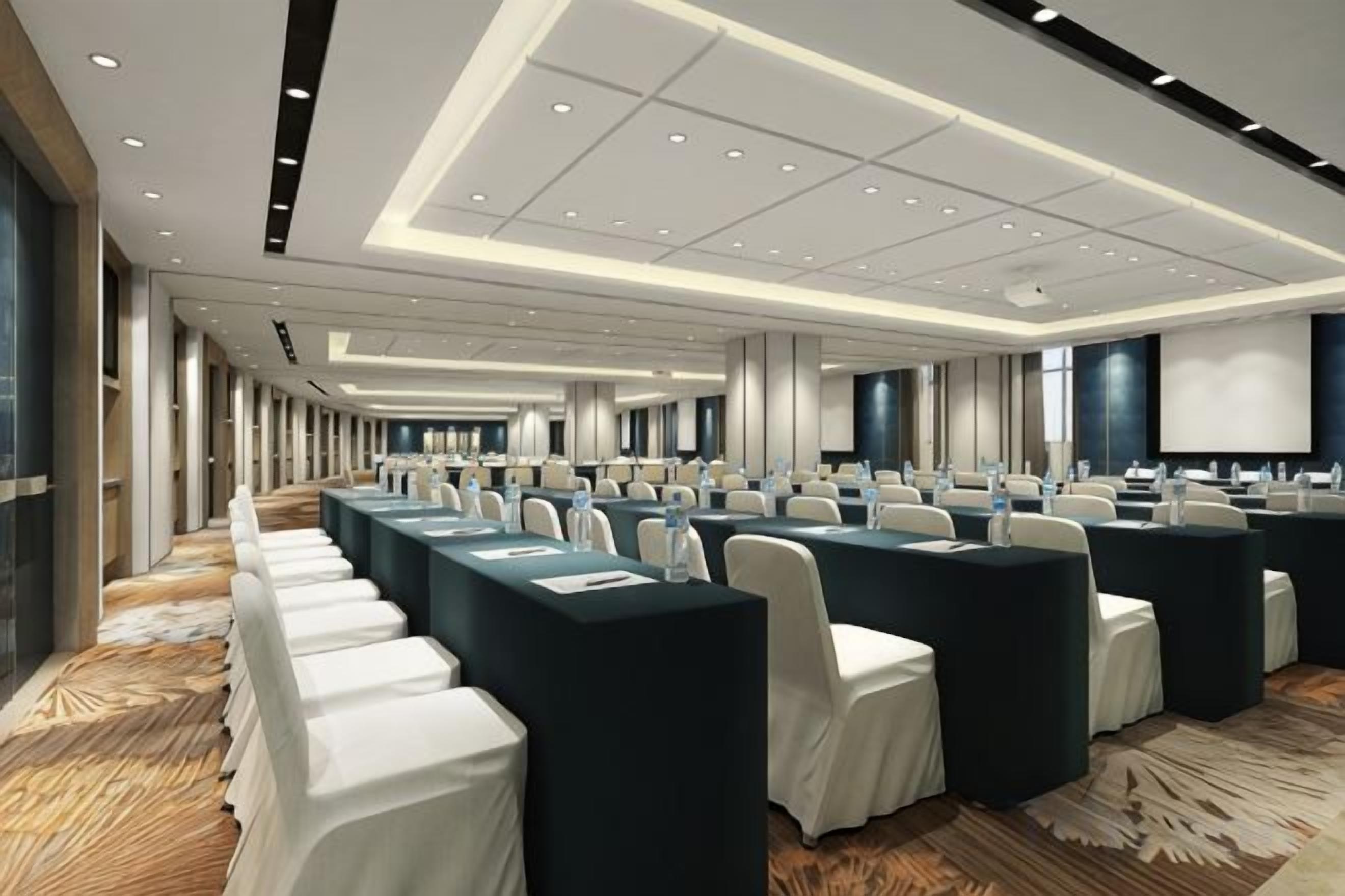 A conference room with rows of tables and chairs, a large screen, and a carpeted floor.