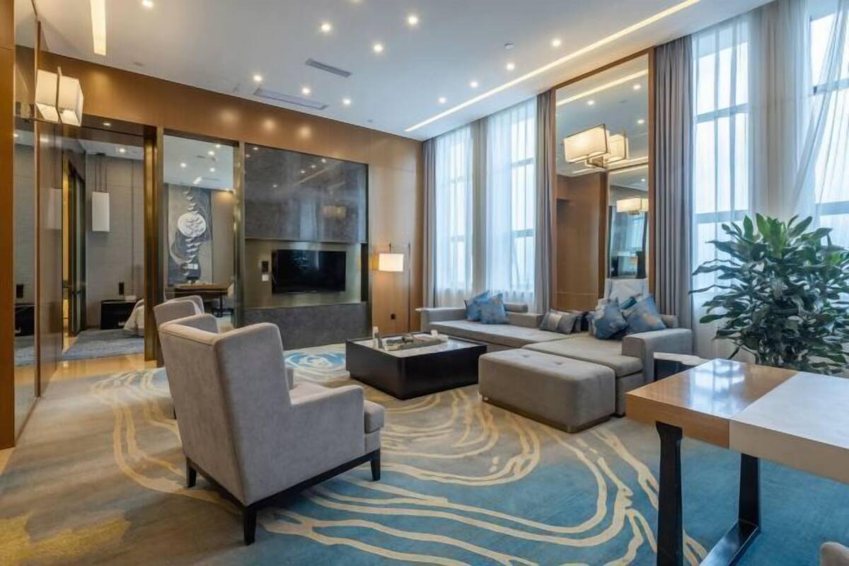 A modern hotel room with a large sofa set, a flat-screen TV, a fireplace, and a large window with curtains.