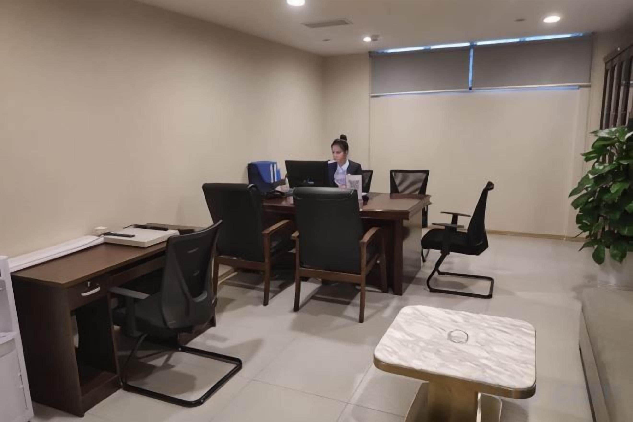 A conference room with a person at a desk, multiple chairs, a table, and a potted plant.