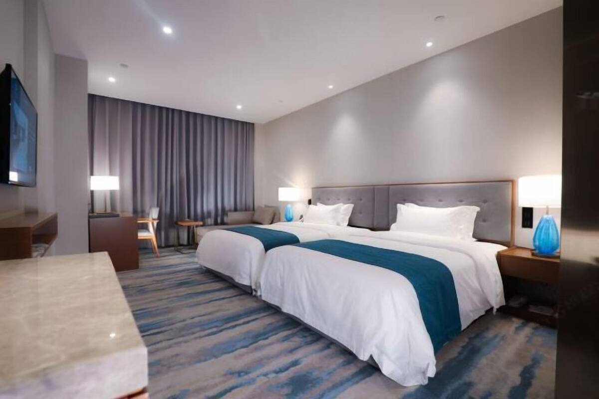 A hotel room with two beds, a desk, a chair, a TV, and a large window with curtains.