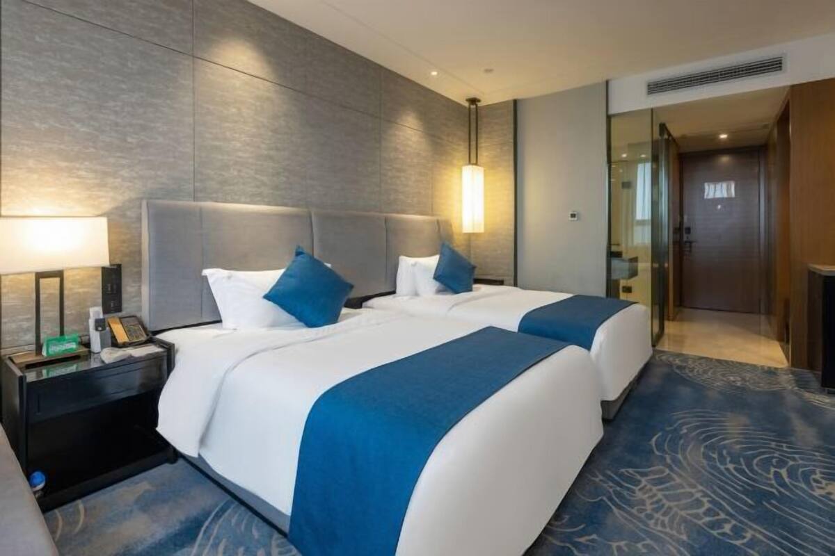 A hotel room with two beds, a nightstand, a lamp, and a large window.