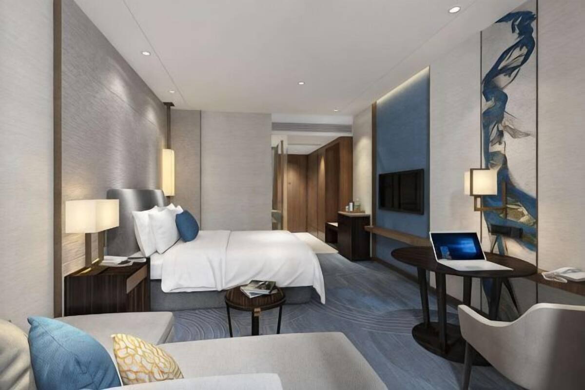 A modern hotel room with a bed, a desk with a laptop, a chair, a sofa, and a TV.